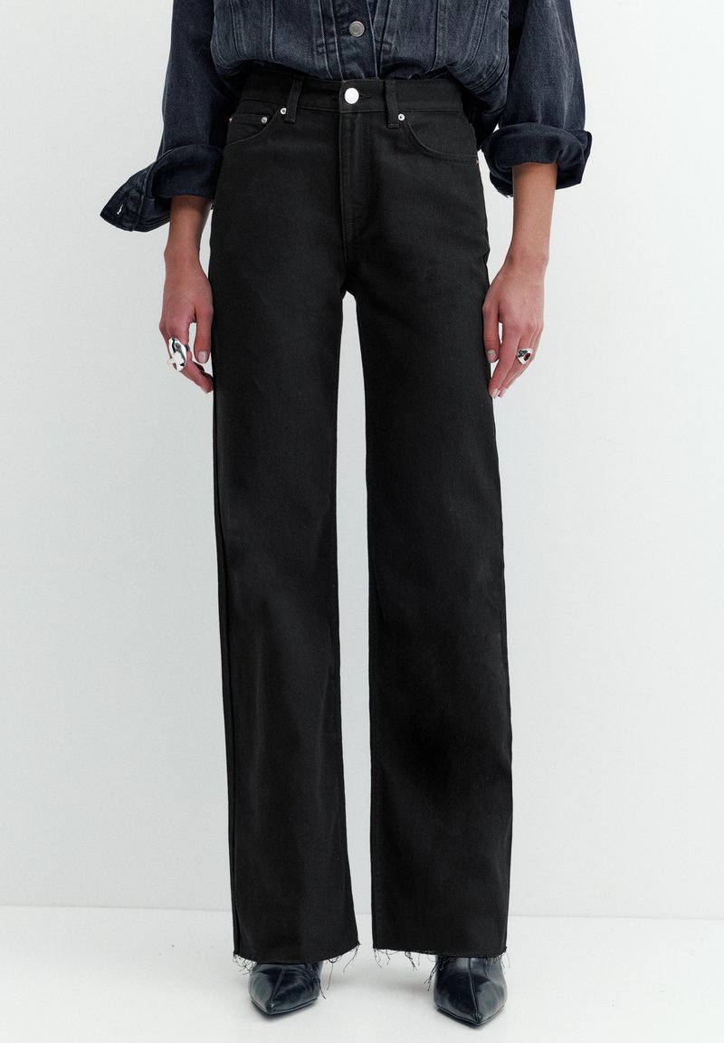 Osoba nosząca czarne dżinsy z wysokim stanem i szerokimi nogawkami, z surowymi wykończeniami, ciemną denimową koszulę z podwiniętymi rękawami oraz czarne, spiczaste buty.