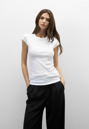 Kvinde med langt brunt hår iført en tætsiddende hvid kortærmet top og løse sorte bukser, stående med hænderne i lommerne mod en ensfarvet baggrund.