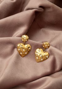 Orecchini a forma di cuore in oro con superfici testurizzate, dotati di gemme trasparenti incastonate, esposti su un morbido tessuto color taupe.