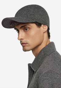 Grå stickad mössa med en böjd skärm, tillverkad av ett texturerat tyg. Mössan är i kombination med ett matchande grått plagg med krage, vilket framhäver en sammanhängande look.