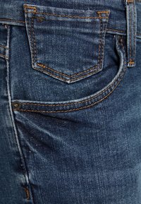 Tmavě modré džíny z denimu, které mají zadní kapsu s oranžovým šitím a malým černým logem nad kapsou, ukazující texturovanou tkaninu.