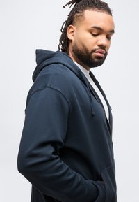 Άνδρας με κράμπλες μαλλιών, φοράει λευκό μπλουζάκι κάτω από μπλε-ναυτικό φούτερ με φερμουάρ, κοιτάζει προς τα κάτω με τα χέρια στις τσέπες, με λευκό απλό φόντο.