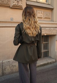 Trench coat verde oliva con cintura e dettaglio a patta, indossato sopra pantaloni grigi larghi, abbinato a una piccola borsa nera a spalla. Sfondo texturizzato.