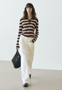 Gestreiftes Langarmoberteil in Braun und Beige, kombiniert mit weißen Hosen mit weitem Bein. Das Modell trägt eine schwarze strukturierte Tasche und schwarze Schuhe.