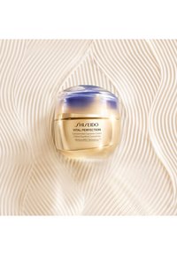 Shiseido Vital Perfection-kräm i en guldig och blå burk, rund form med en slät textur, visad mot en vågig krämfärgad bakgrund.