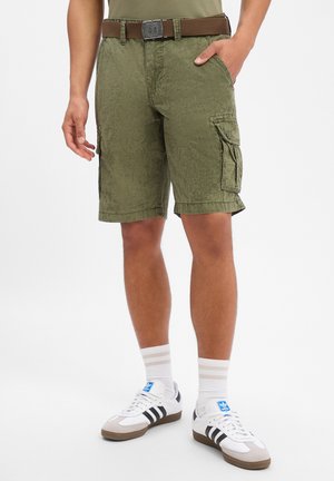 Mann trägt olivgrüne Cargo-Shorts mit braunem Gürtel, weißen Adidas-Sneakern mit schwarzen Streifen und weißen Sportsocken mit beigen Streifen.