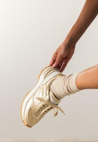 Zapatilla deportiva dorada con superficie texturizada, media suela blanca y suela de goma color marrón. Presenta cordones a juego y un diseño que llega hasta el tobillo.