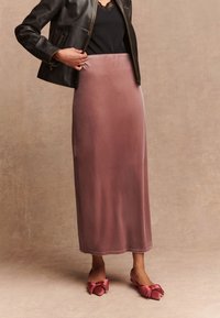 Langer, rosafarbener Samtrock kombiniert mit einem schwarzen ärmellosen Oberteil und einer taillierten schwarzen Lederjacke, ergänzt durch Schuhe mit rosa Schleifendetail.