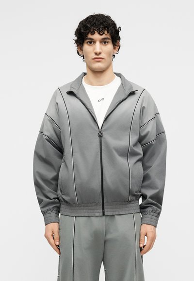 Νέος με σγουρά μαλλιά, φοράει γκρι ζακέτα με φερμουάρ και αντίστοιχο παντελόνι με λευκές λεπτομέρειες, στέκεται μπροστά σε ένα απλό φόντο.