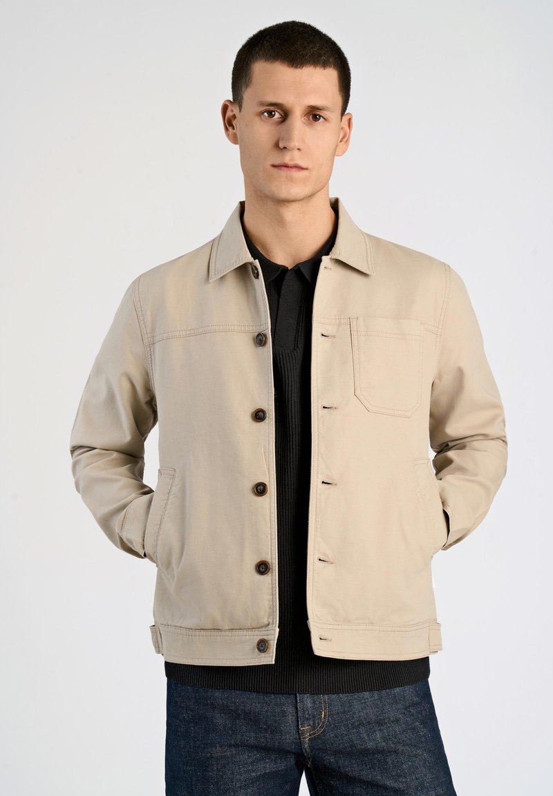 Giacca di cotone beige con colletto, bottoni frontali e tasche laterali; indossata sopra un maglione nero con texture e jeans di denim blu.