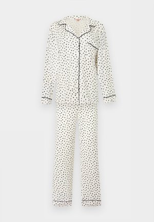 Nő áll mezítláb, fehér, hosszú ujjú, gombos pizsamát visel fekete szív mintákkal és fekete piping részletekkel.