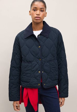 Femme portant une veste matelassée bleu marine avec grand col, poches avant et boutons-pression en laiton sur un haut blanc et un vêtement rouge noué autour de la taille.