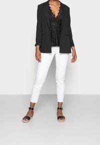 Donna con top di pizzo nero, blazer nero, jeans bianchi a vita alta e sandali neri con cinturino alla caviglia, in piedi davanti a uno sfondo neutro.