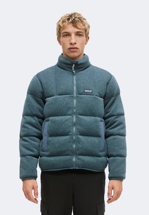 Junger Mann trägt eine blaue Steppjacke mit Frontreißverschluss und zwei Seitentaschen vor einem schlichten weißen Hintergrund.