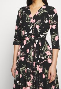 Blumenkleid in schwarzem Stoff mit rosa- und weißen Blumen, mit Dreiviertelärmeln, einem V-Ausschnitt und einem Taillenzug für den Verschluss.