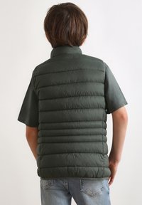 Grüne Steppweste mit gepolstertem Design, hohem Kragen und kurzen Ärmeln, getragen über einem dunklen Shirt, kombiniert mit hellblauen Jeans.