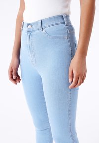 Vaqueros de denim de color azul claro con cintura alta, corte slim, desvanecido sutil y cierre estándar de botón y cremallera. Costuras visibles a lo largo de las costuras laterales.