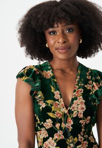 Green floral dress with ruffled sleeves, featuring a deep V-neckline, velvet texture, and pink and yellow flower patterns. Gold earrings accentuate the look.