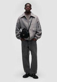 Šedý kabát z vlněné směsi s velkými kapsami, vrstvený přes hnědou košili, spárovaný s tmavě šedými džíny a černými botami na slip-on. Součástí je černá kabelka.