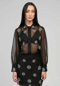 Blouse noire transparente à manches longues, devant boutonné, avec broderies florales en accents argentés et dorés, assortie à une jupe brodée.