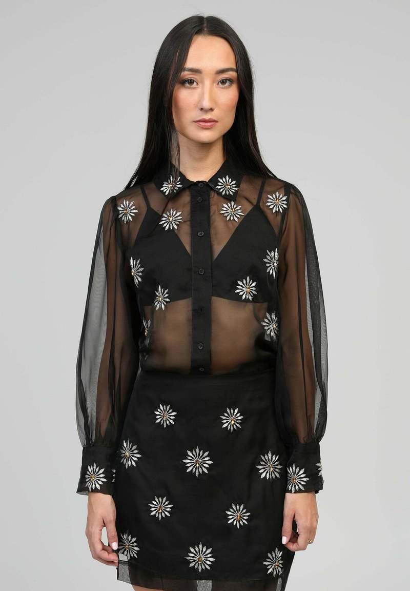 Blouse noire transparente à manches longues, devant boutonné, avec broderies florales en accents argentés et dorés, assortie à une jupe brodée.