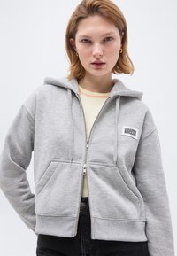 Νεαρή γυναίκα με κοντά ξανθά μαλλιά, που φορά χρυσούς κρίκους στα αυτιά, ανοιχτογκρι ζακέτα με φερμουάρ και ανοιχτόχρωμο πουκάμισο, στέκεται μπροστά σε απλό φόντο.