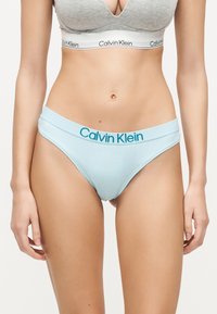 Femme portant un slip bikini bleu clair Calvin Klein et un soutien-gorge de sport gris Calvin Klein, debout devant un fond blanc uni.