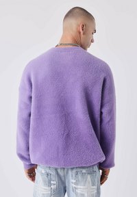 Suéter morado esponjoso con mangas largas y un dobladillo redondeado. Presenta una textura suave y un diseño relajado, combinado con jeans de mezclilla clara.