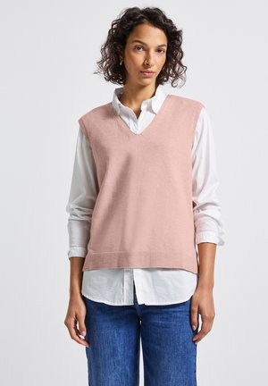 Femme aux cheveux bouclés portant un pull sans manches V en rose clair sur une chemise blanche et un jean bleu, se tenant devant un fond uni.