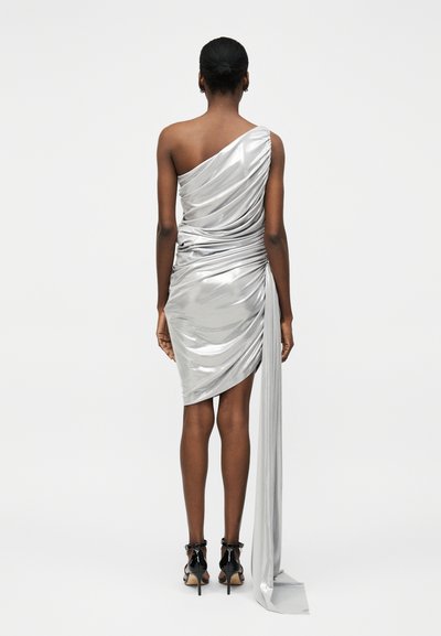 Γυναίκα στέκεται με την πλάτη, φοράει ένα γυαλιστερό ασημί φόρεμα με έναν ώμο και ρυτιδώσεις, μακριά υφάσματα και μαύρα πέδιλα με ψηλό τακούνι.