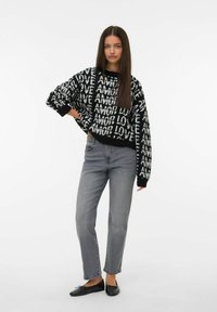 Giovane donna con lunghi capelli scuri, che indossa un maglione a righe nere e bianche con la scritta "LOVE AMOR", jeans grigi e ballerine nere, in piedi con una mano sulla vita.
