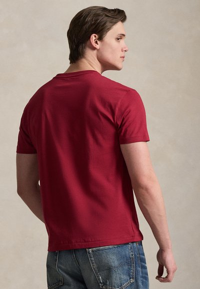 Νέος άνδρας σε πλάγια όψη, φορώντας απλό κόκκινο κοντομάνικο μπλουζάκι και μπλε τζιν παντελόνι, μπροστά σε ουδέτερο φόντο.