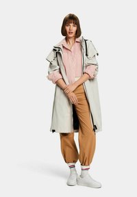Cappotto trench grigio chiaro con vestibilità rilassata, indossato sopra una maglietta a righe rossi e bianchi, abbinato a pantaloni jogger beige e sneaker bianche.