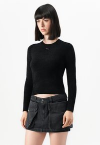 Donna con capelli lunghi e lisci di colore scuro, che indossa un top aderente nero a maniche lunghe e una mini gonna in denim nero con ampie tasche frontali.