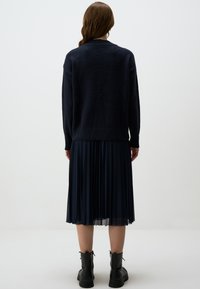 Pull en maille bleu marine à coupe décontractée, superposé à une jupe plissée bleu marine, associé à des bottines noires, vu de dos.
