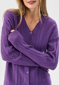 Gilet en tricot torsadé violet avec un col en V, présentant des poignets côtelés et une fermeture par bouton central. Texture douce et détails en motif de diamants.