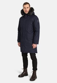 Parka navy con cappuccio foderato in pelliccia, tasche anteriori, tasca sul petto con zip e taglio dritto, indossata con pantaloni neri e stivali marroni.