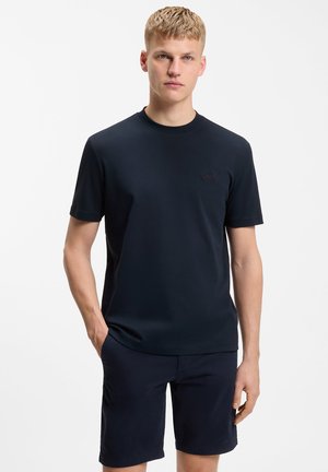 Νεαρός άνδρας στέκεται με τα χέρια στις τσέπες, φορώντας απλό σκούρο μπλε κοντομάνικο πουκάμισο και αντίστοιχα σορτς, μπροστά σε λευκό φόντο.