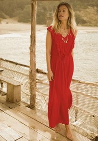Red maxi dress with ruffle accents, tie waist, and a flowing skirt. Worn barefoot against a natural, outdoor backdrop.