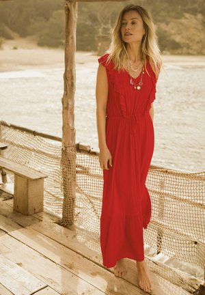 Red maxi dress with ruffle accents, tie waist, and a flowing skirt. Worn barefoot against a natural, outdoor backdrop.