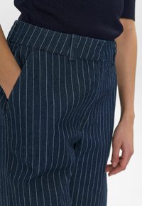 Jūriniai dryžuoti kelniai iš tekstūruotos medžiagos, turintys juosmenį, šonines kišenes ir laisvą siluetą. Vertikalios baltos juostelės pabrėžia dizainą.