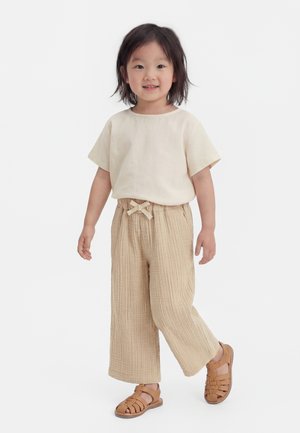 Young child wearing a cream short-sleeve shirt, beige wide-leg pants with a bow, and brown closed-toe sandals, standing on a white background.