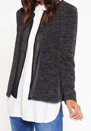 Femme portant un cardigan gris foncé ouvert sur une chemise blanche boutonnée, associée à un jean bleu.