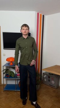 Νέος άνδρας με τατουάζ στα χέρια, φοράει πράσινο πουκάμισο και σκούρο παντελόνι, στέκεται σε ένα δωμάτιο με τηλεόραση, πορτοκαλί λάμπα, φυτά και πολύχρωμες ρίγες στον τοίχο.