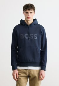 Νέος άνδρας με σγουρά μαλλιά, που φοράει μια μπλε σκουρόχρωμη μπλούζα με κουκούλα και την επιγραφή "BOSS" και μπεζ παντελόνι, στέκεται μπροστά σε ένα απλό λευκό φόντο.