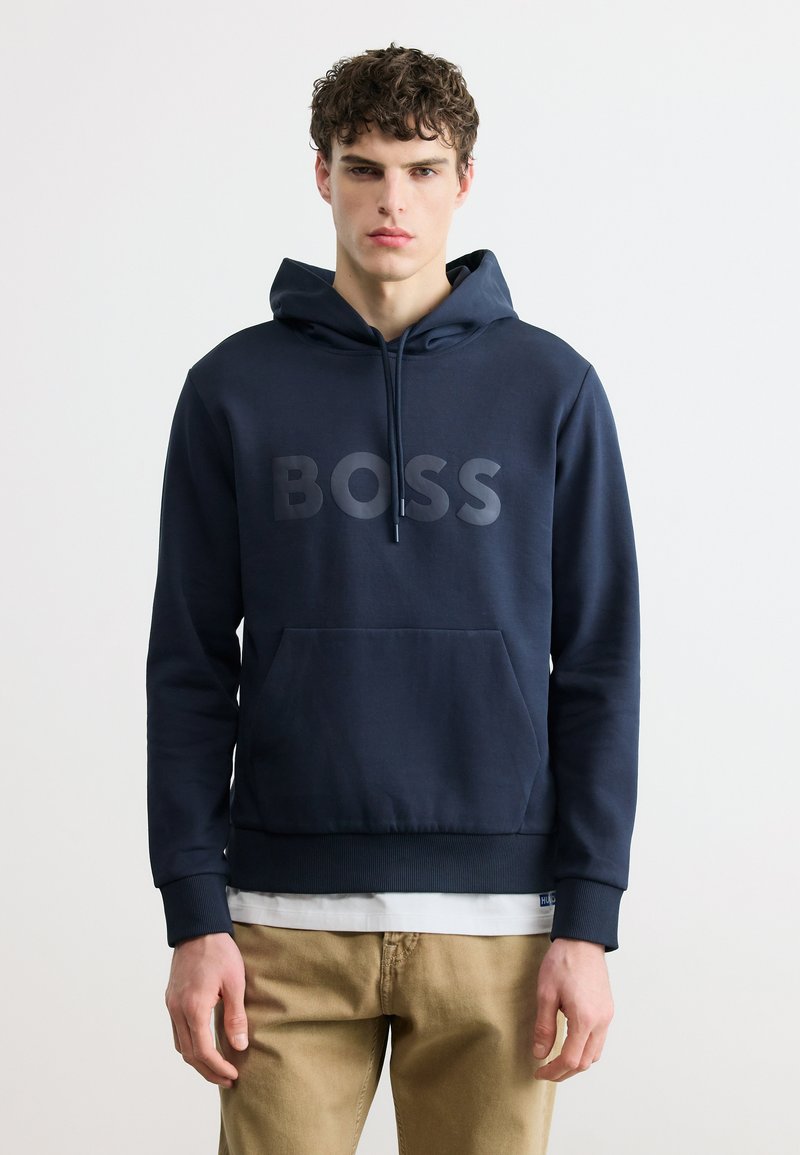 Νέος άνδρας με σγουρά μαλλιά, που φοράει μια μπλε σκουρόχρωμη μπλούζα με κουκούλα και την επιγραφή "BOSS" και μπεζ παντελόνι, στέκεται μπροστά σε ένα απλό λευκό φόντο.