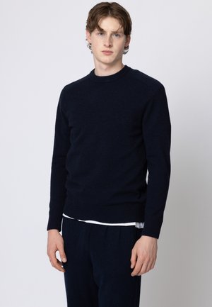 Giovane uomo con pelle chiara e capelli castani mossi che indossa un maglione blu navy scuro e pantaloni coordinati su uno sfondo chiaro e uniforme.