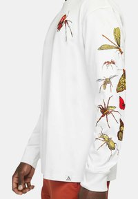Långärmad vit skjorta med färgglada insektsgrafik längs ärmarna. Har en etikett vid fållen med en triangulär logotyp.