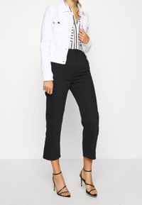 Veste en jean blanche sur une chemise à rayures noires et blanches, associée à un pantalon noir taille haute et court, et des talons noirs à brides.