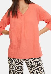 Blusa in lino corallo con scollatura a V, maniche a tre quarti e vestibilità comoda, abbinata a pantaloni a pattern bianco e nero.